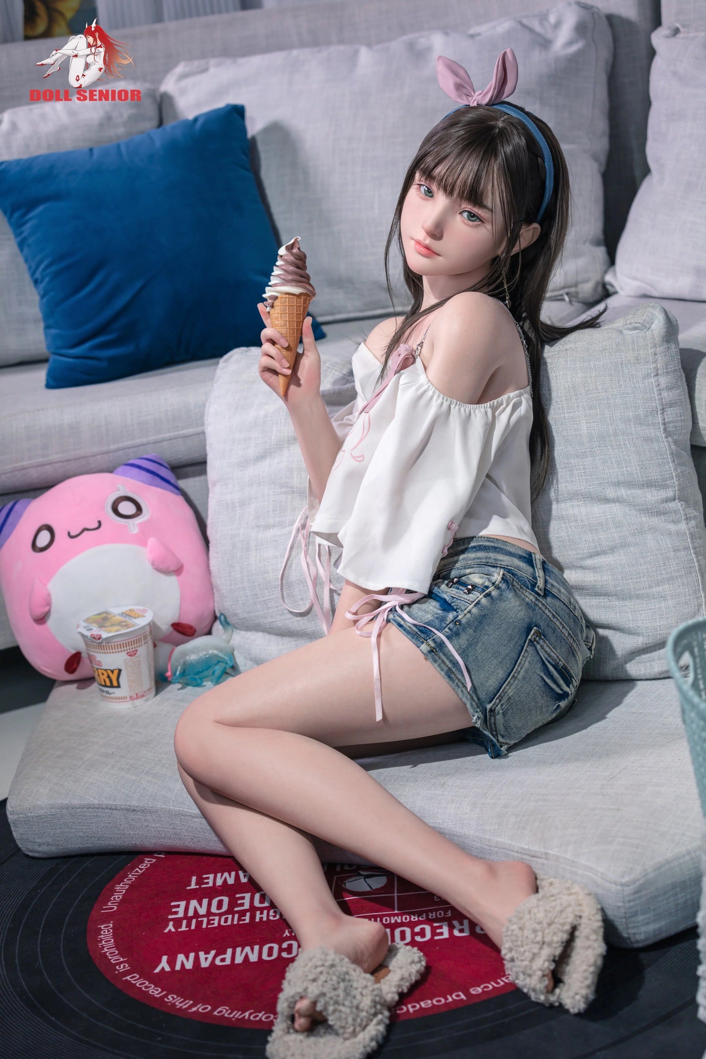 Realistic female doll with brown hair, bunny ears, holding ice cream, wearing white top and denim shorts on gray couch