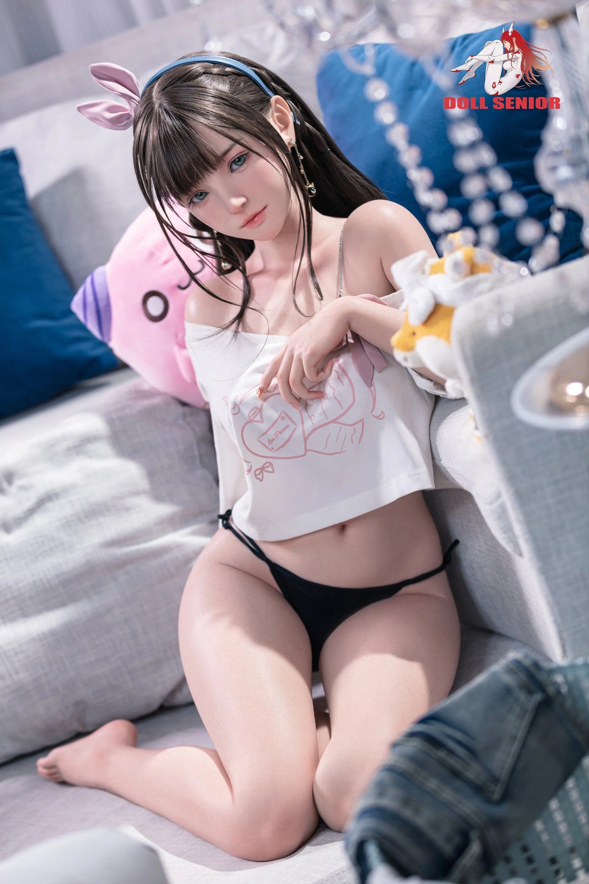 Realistic female doll with brown hair, blue eyes, wearing white crop top and black lingerie, sitting on couch with plush toys.