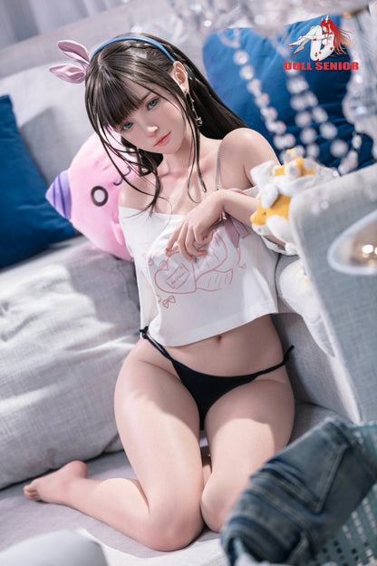 Realistic female doll with brown hair, blue eyes, wearing white crop top and black lingerie, sitting on couch with plush toys.