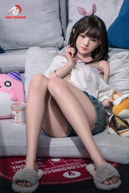 Realistic full-body doll with brown hair, green eyes, wearing white top, denim shorts, and bunny ears, seated on gray sofa with plush toys.