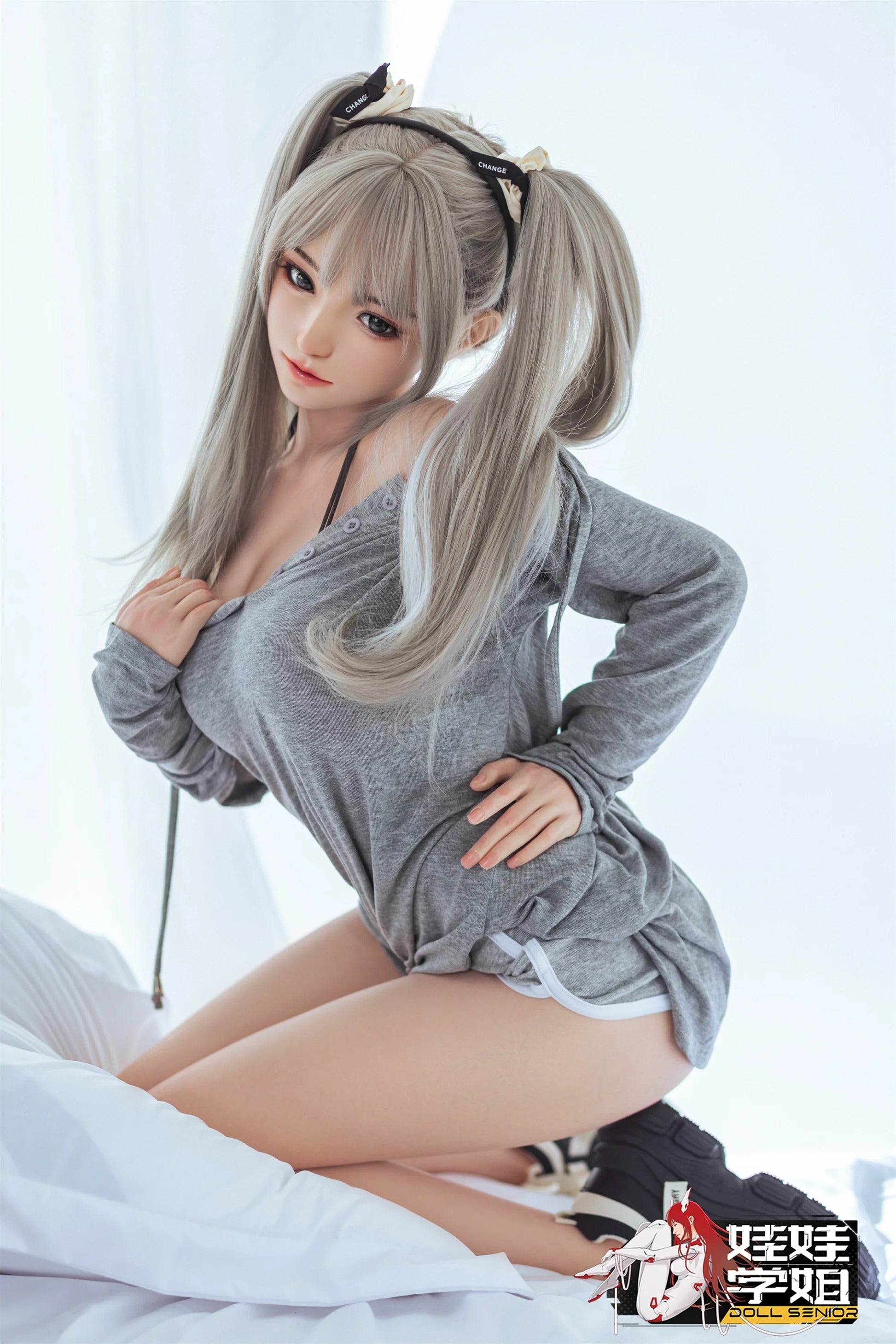 Realistic female doll with silver pigtails, wearing gray loungewear, posed on white bedding.