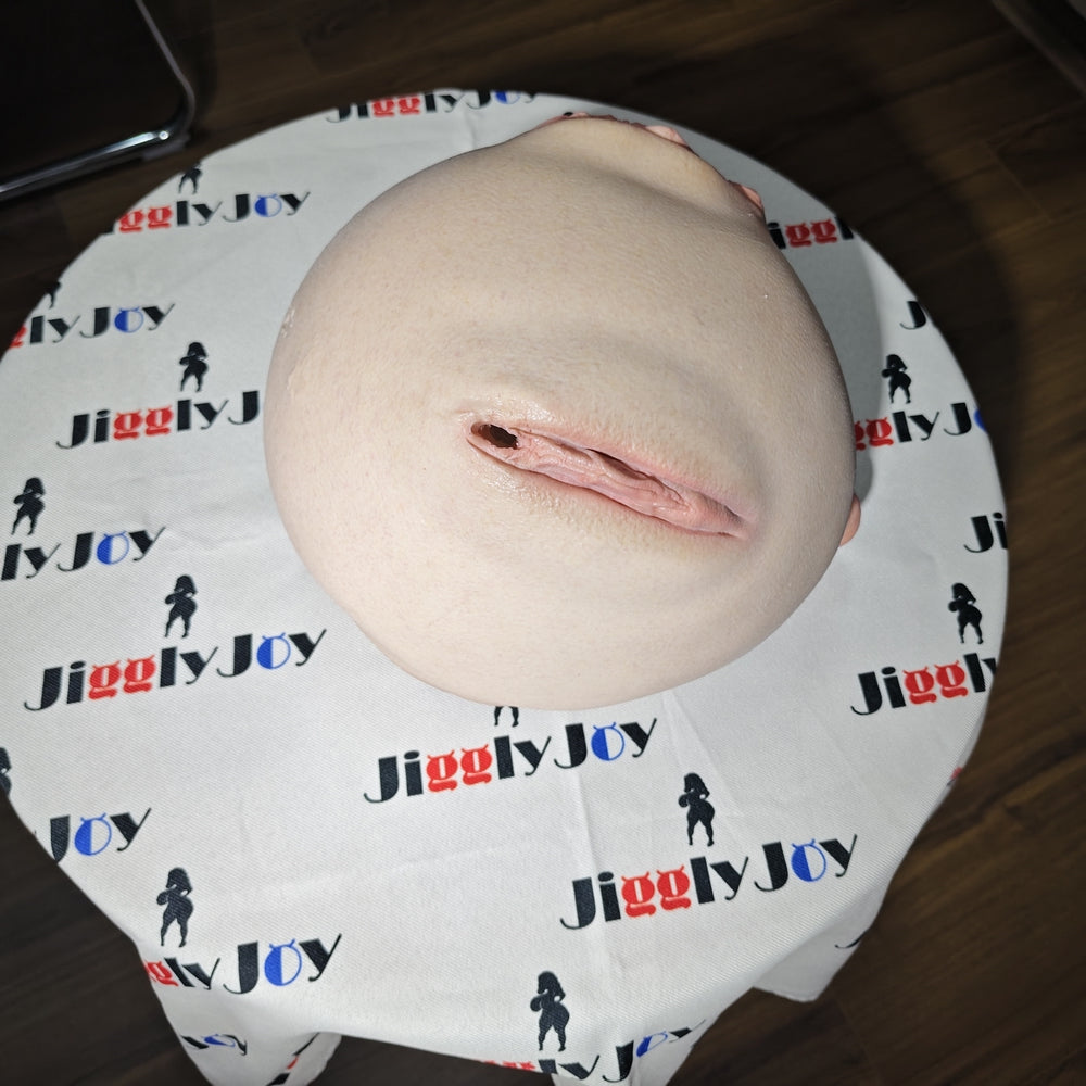 Realistic flesh-toned male masturbator with detailed vaginal opening, resting on JigglyJoy-branded fabric